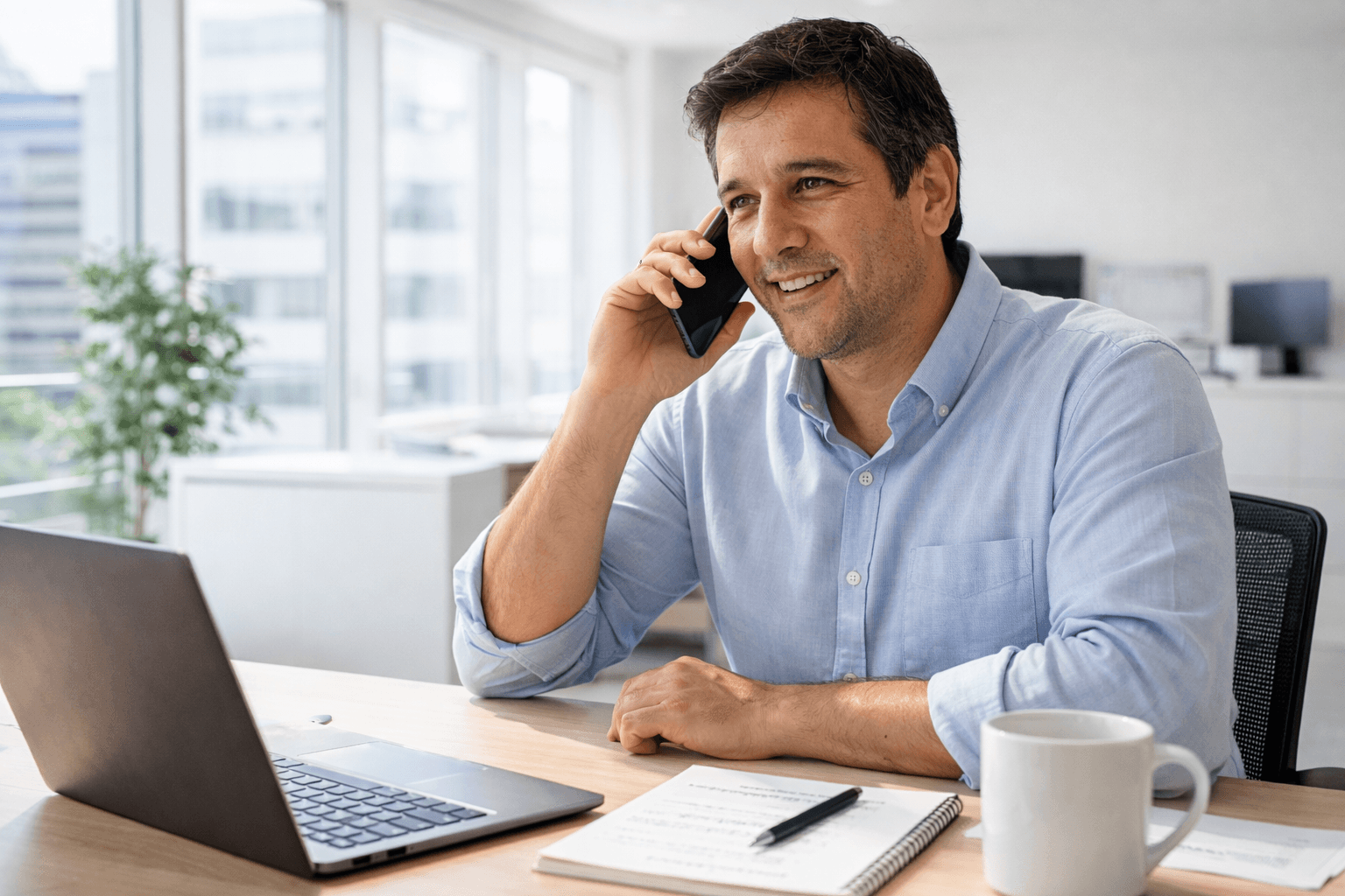 Smiling man talking on a smartphone at an office desk with a laptop and notebook.